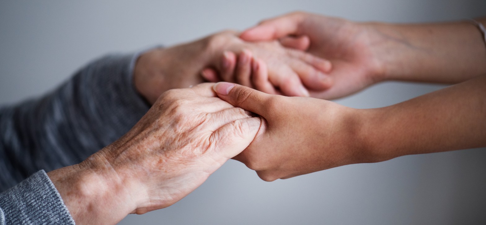 A la izquierda las manos de una persona mayor son sujetadas en actitud de ayuda y apoyo por las manos de una persona joven a la derecha de la imagen.