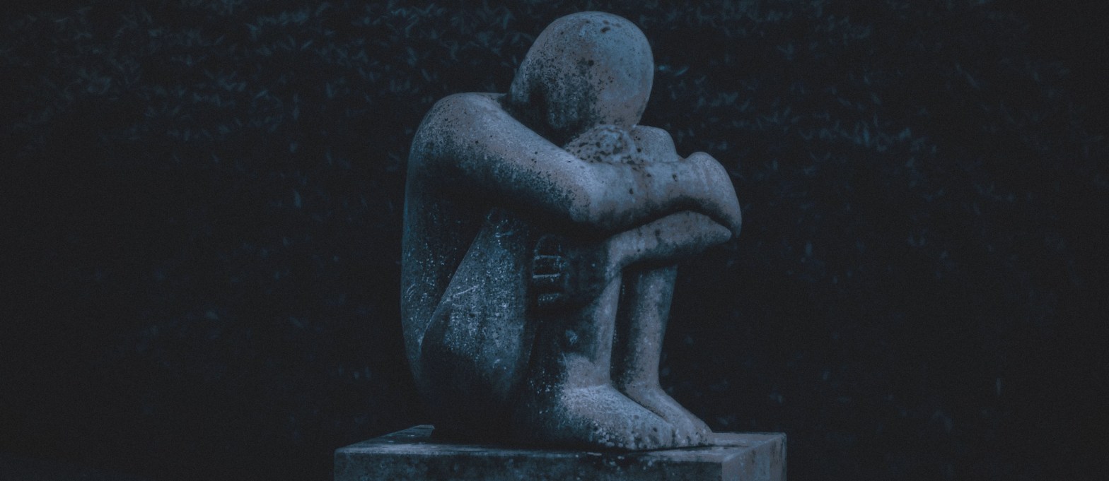 La imagen muestra una estatua de piedra en la que se ve una figura humana sentada cogiéndose las piernas con los brazos formando una postura que inspira la depresión o la tristeza.