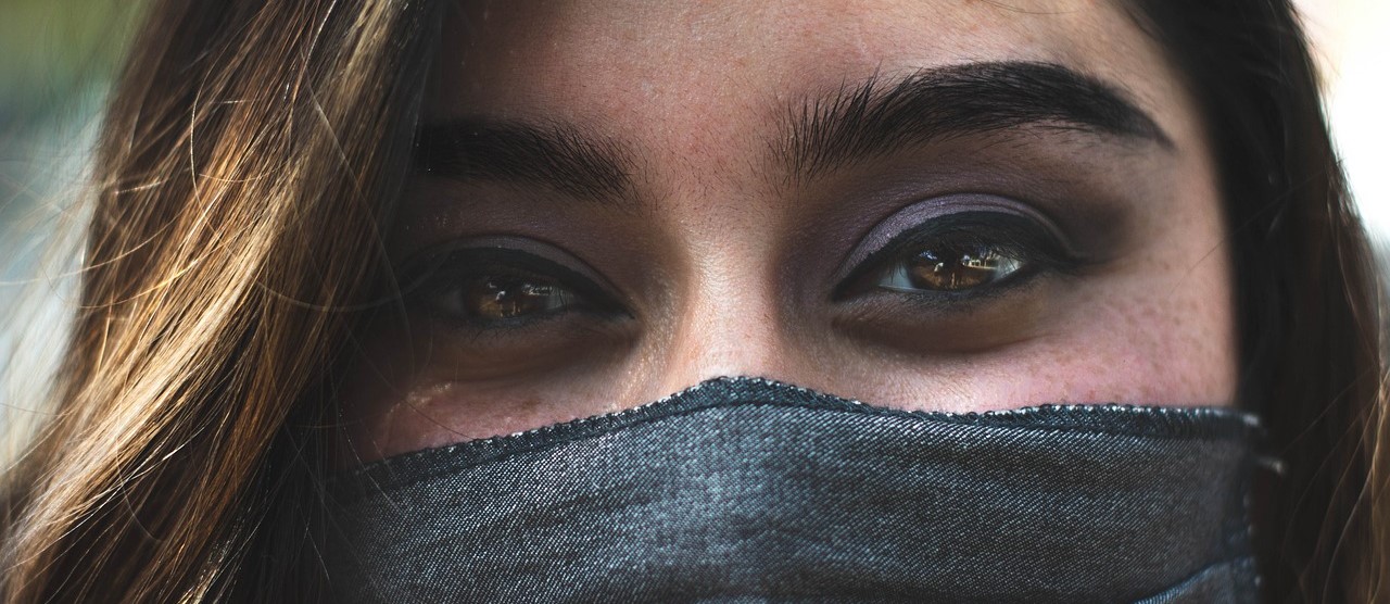 La imagen es un primer plano muy cerrado del rostro de una mujer de tez morena, pelo y ojos castaños, que lleva una mascarilla de tela que le tapa la parte inferior del rostro desde la nariz.