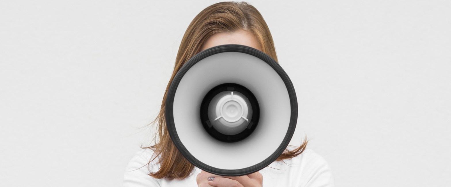 Una persona con el pelo largo hasta los hombros empuña un megáfono con las dos manos apuntando directamente hacia la cámara.