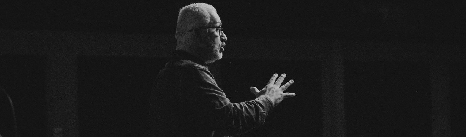 Fotografía en blanco y negro. En el centro hay un hombre de mediana edad de perfil con la mano derecha medio extendida y completamente abierta.